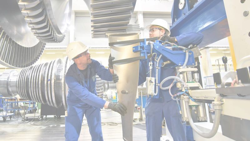 Zwei blau gekleidete Facharbeiter mit weißen Helmen arbeiten an einer Turbine in einer Montagehalle