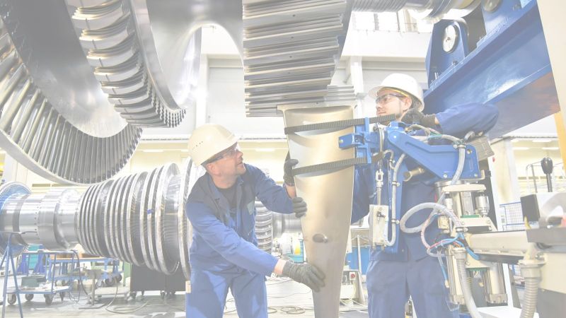 Zwei blau gekleidete Facharbeiter mit weißen Helmen arbeiten an einer Turbine in einer Montagehalle
