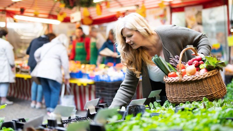 Ein Frau beugt sich suchend an einem Stand auf dem Wochenmarkt über die angebotenen Waren. In der einen Hand hat sie einen Korb voller Gemüse .