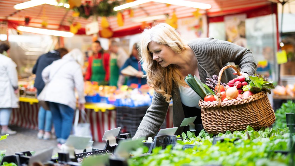 Ein Frau beugt sich suchend an einem Stand auf dem Wochenmarkt über die angebotenen Waren. In der einen Hand hat sie einem Korb voller Gemüse .